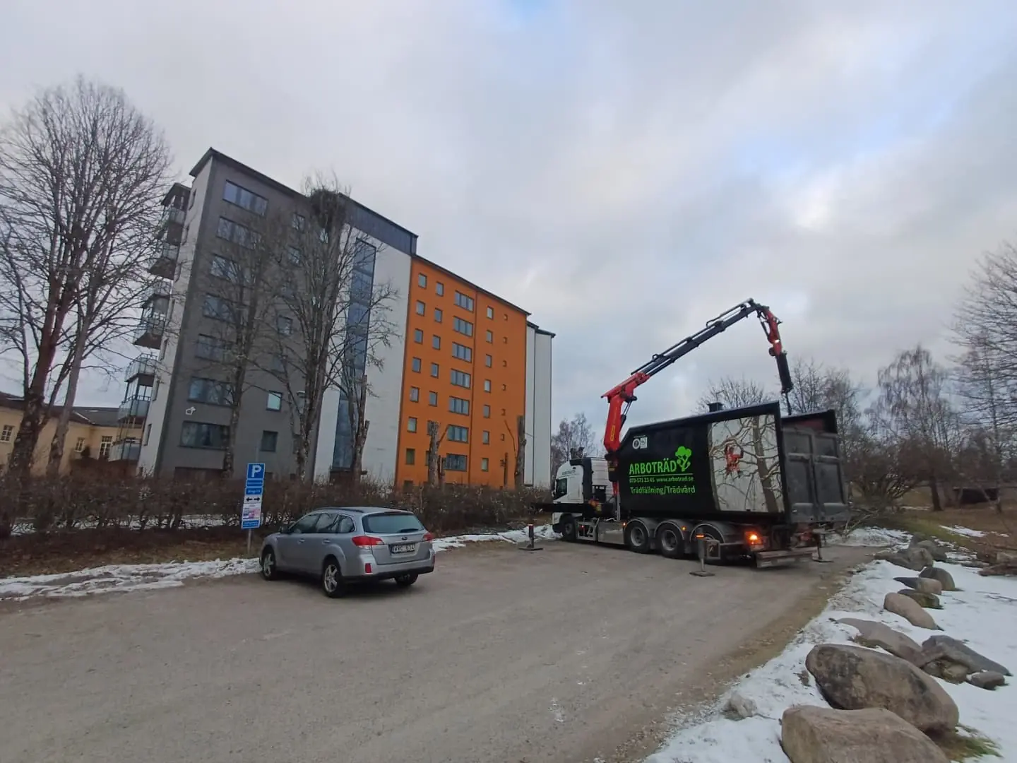 Arboträds kranbil vid flerbostadshus en vinterdag, kranen uppfälld mot ett träd